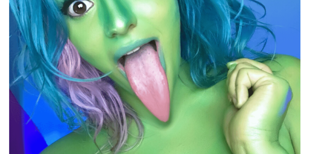Selfie of Jenn sticking out her tongue. Her skin is tinted green and her tongue is edited so it's extra long. She has blue, green, and pink hair and her left hand is up by her shoulder.